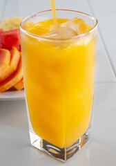 Serving a mango juice in a sqaure shaped glass with ice, on a white kitchen surface and a plate of sliced fruits on the background. Vertical image