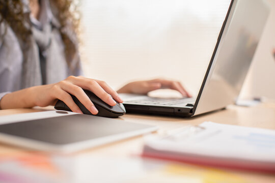 Close up hand of business asian young woman with curly hairstyle holding digital computer mouse and clicking working with computer laptop.Female freelancer using wireless mouse on desk at home