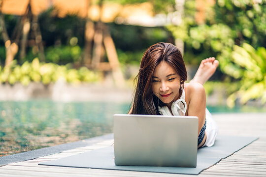 Portrait of smiling happy beauty asian freelancer traveler blogger woman relax using and work technology of laptop computer in swimming pool on summer travel vacation relaxing at resort spa