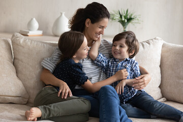 Happy mom hugging little son and daughter with love, care, tenderness, relaxing with kids in couch,...