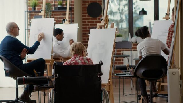 Old woman with disability using pencil to draw vase artwork, attending creative lesson in wheelchair to develop new drawing skills. Educational activity in workshop. Tripod shot.
