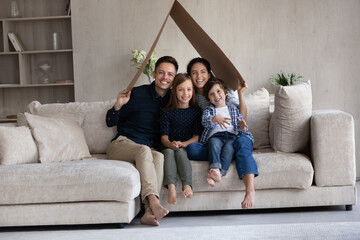 Happy young parents and two cute kids holding cardboard paper roof of house overhead, showing...