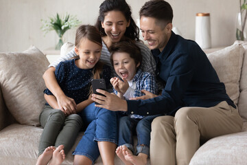 Happy young parents and excited kids watching funny video online content on mobile phone, making call, talking, using app, getting good news, feeling joy. Family using smartphone, relaxing on couch