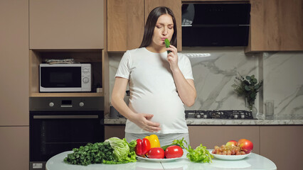 Pregnant woman with long brunette hair wearing white t-shirt and training pants eats vegetables put on table stroking large belly in kitchen.