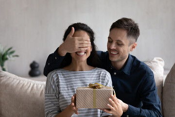 Happy husband giving surprise gift to excited wife. Joyful young man covering girlfriends eyes,...