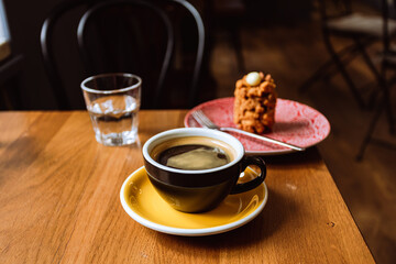 A ceramic cup of black coffee,a dessert served on a plate and a glass of water on the wooden table in a cafe.