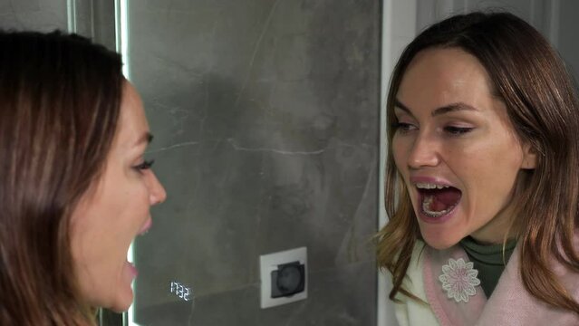 Brunette woman examines dental braces on teeth opening mouth widely and looking in mirror in bathroom against grey tile at home side view, closeup