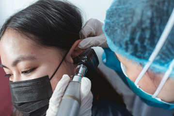 A doctor in full PPE gear checks the ear drum of a female patient with an otoscope, also known as...