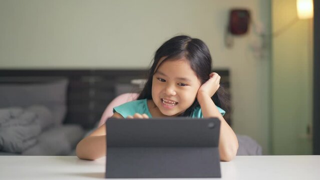 Asian child happy smile talking fun to teacher via video call learning on computer tablet or kid girl student enjoy touch screen and studying online class or person learn from home school in bedroom