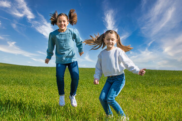 Two Little Girls Enjoying And Jumping On A Green Meadow With A Blue Sky On A Sunny Spring Day. One Has Blonde Hair And The Other Has Brown Hair And They Have Pigtails. Image With Copy Space.
