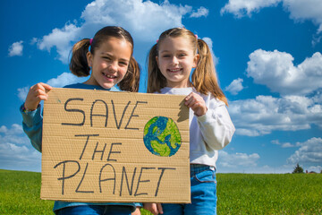 Two Little Girls Holding A Cardboard Sign That Says Save The Planet. They Are In A Green Meadow...