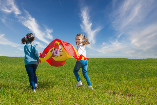 Two Beautiful Girls Running Across A Green Meadow With A Blue Sky, They Are Enjoying And Showing The Flag Of Spain. One Is Blonde And The Other Is Brunette And They Have Pigtails.Image With Copy Space