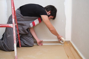 Professional painter using painters tape to cover the skirting board and edges before he starts...
