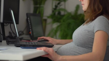 A pregnant woman is working on a laptop. The hand is on the stomach. Pregnant businesswoman typing on a laptop while sitting at her workplace in the office. Work during pregnancy from home.