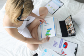 Woman with handsfree sitting on white bed with documents in hands and communicating via video link...