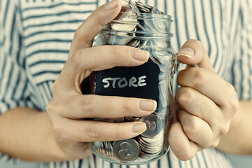 Girl holds glass jar with coins. Black sticker with inscription store on bottle. Savings for purchases. Woman in striped shirt.