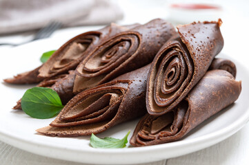 Rolled chocolate crepes laid out in a pile and fresh mint leaves on a white plate. Close-up. Tasty breakfast