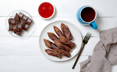 Thin chocolate pancakes rolls with tea and jam on a white wooden background. Top view, flat lay.