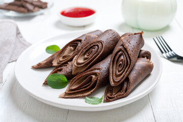 Thin chocolate pancakes rolls with milk and jam on a white wooden background. Tasty breakfast