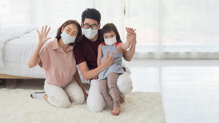 Happy Asian family, mother, father and young disabled child, down syndrome or trisomy 21 daughter putting medical face mask sitting on the floor in bedroom at home during pandemic outbreak of Covid-19
