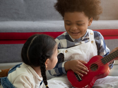 Two adorable diverse children, girl lying down while little Black boy playing Ukulele to girl. Love and happiness, Valentine's concept.