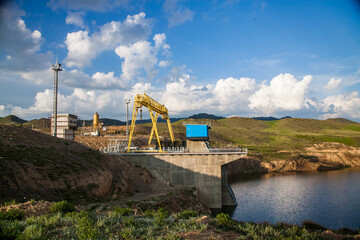 Dam on Sharyn river. Artificial lake. Blue machine house, yellow gantry crane. Green mountains....