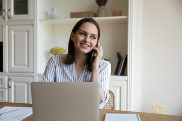 Distracted from working on laptop happy young businesswoman listening pleasant news enjoying cellphone call conversation, discussing online project success with colleagues, multitasking in office.