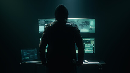 Back view of unrecognizable man in hoodie standing near desk and reading stolen data from computer,...