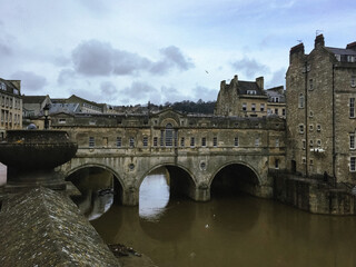 Fototapeta premium Pulteney bridge over Avon river in Bath. Somerset, England. Old town, stone walls, picturesque spot, blue sky, flying bird, dark water. Chimney. Rapids on a river. Stream. Moss
