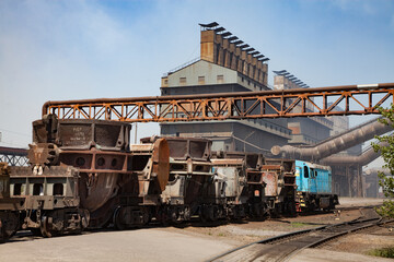 Metal alloys plant. Rusted slag cars and blue locomotive on rails. Metallurgical plant (smelter) main industrial building with chimneys on background.