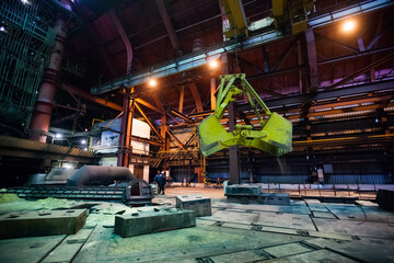 Arcelor Mittal metallurgy plant. Steel manufacturing. Smelter interior. Green mechanical slag hand. Electric arc furnace left. Two workers down. Temirtau, Kazakhstan