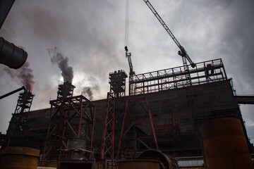 Silhouette  view on modern smelter. Industrial building, smoke stacks with smoke and crane. Dark grey cloudy sky.