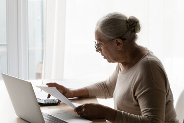 Senior 70s landlady, tenant lady using calculator, online payment banking app on laptop, checking paper bills, paying taxes, insurance fees, calculating taxes, money, budget, doing paperwork