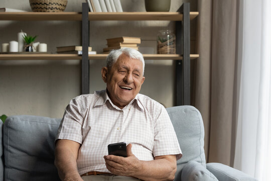 Happy joyful older 80s man holding, using online ecommerce app on mobile phone, resting on sofa at home, chatting on internet, laughing, making call. Distant communication concept