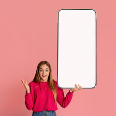 Check out this new mobile app. Excited woman presenting smartphone with white empty screen over pink background - obrazy, fototapety, plakaty