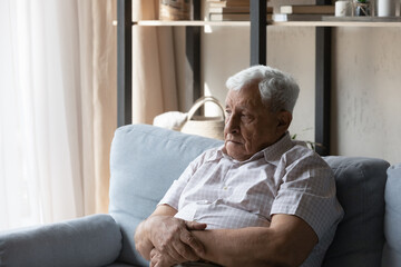 Frustrated senior elder 80s man sitting on sofa at home, looking away, missing family, feeling...