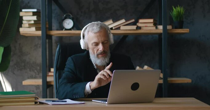 Happy bearded mature businessman listening music in headphones while working at laptop computer at office