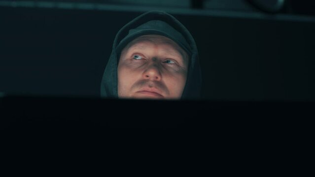 Low angle pan left view of serious male hacker in hoodie and cap reading data on computer monitors, while hacking database of big data servers in darkness.