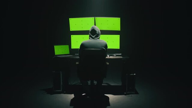 Rear view of hooded anonymous scammer hacker at table with green screen chromakey multiple display, during an attack on credit cards big data servers on the network