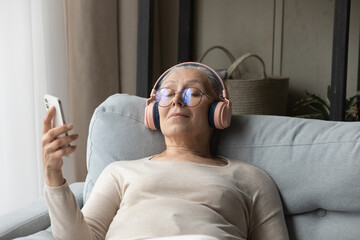 Calm peaceful senior lady in big wireless headphones and glasses listening to relaxing music from...