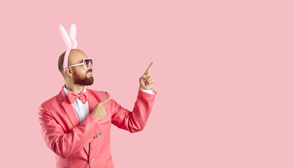 Funny young man wearing easter bunny ears shows on copy space on pastel pink background. Cheerful...