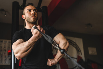 One man young adult caucasian male bodybuilder training arms bicep on the cable machine in the gym holding weight wearing black shirt dark photo real people copy space side view low angle