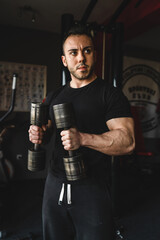Fototapeta na wymiar One man young adult caucasian male bodybuilder training arms bicep flexing muscles with dumbbell while standing in the gym wearing black shirt dark photo real people copy space front view waist up