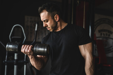 One man young adult caucasian male bodybuilder training arms bicep flexing muscles with dumbbell while standing in the gym wearing black shirt dark photo real people copy space front view waist up