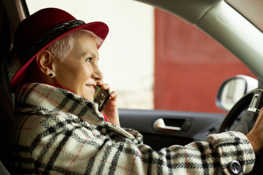 Close-up of elegant fifty-years-old woman in red hat sitting in parked car talking on phone, smiling, having nice pleasant conversation, pictured in profile, wearing plaid stylish trench coat