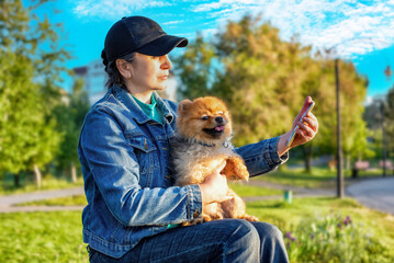 A woman sitting on a bench in the park dialing a smart phone with a Pomeranian dog in summer.