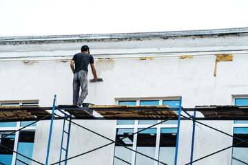 Restoration and repair of the facade of the building. Plasterer works standing on scaffolding without insurance. The process of puttying the wall of a low house with large windows.