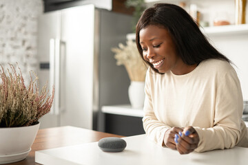 Smiling African-American woman is using smart home assistant device with voice recognize function, multiracial lady talking to portable smart speaker. Smart home concept