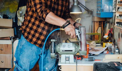 Fototapeta premium An Anonymous Carpenter Working with Bench Grinder in his Workshop. Unrecognizable man cleaning his electric sander with air compressor at his garage.
