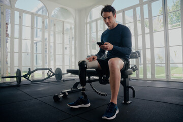 Paralympic athlete on his mobile in the gym training. Man with prosthesis sitting on bench looking...